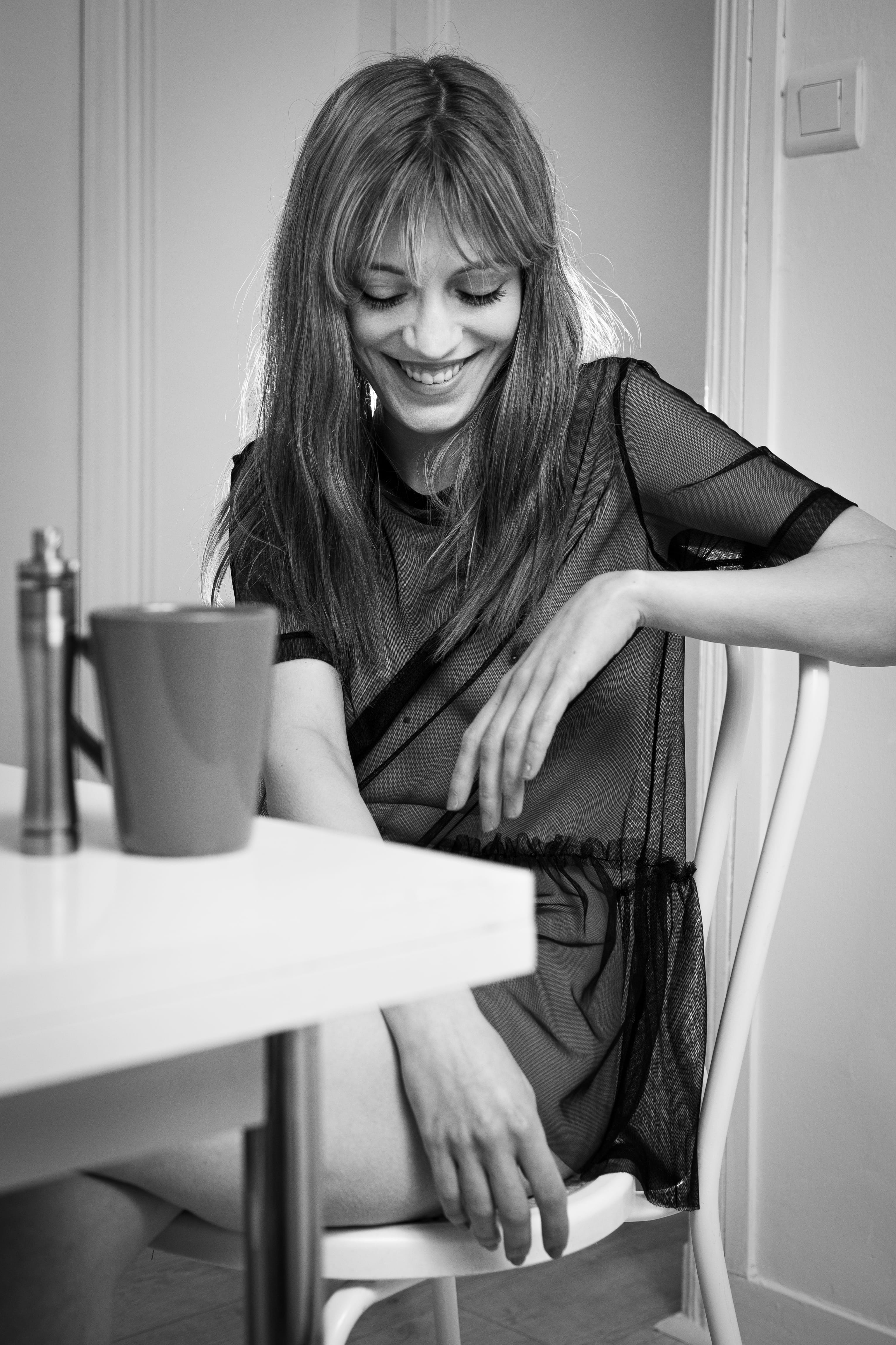Femme souriante assise à une table blanche, vêtue d'une tenue noire transparente, capturée en noir et blanc doux pour une ambiance boudoir détendue et naturelle.