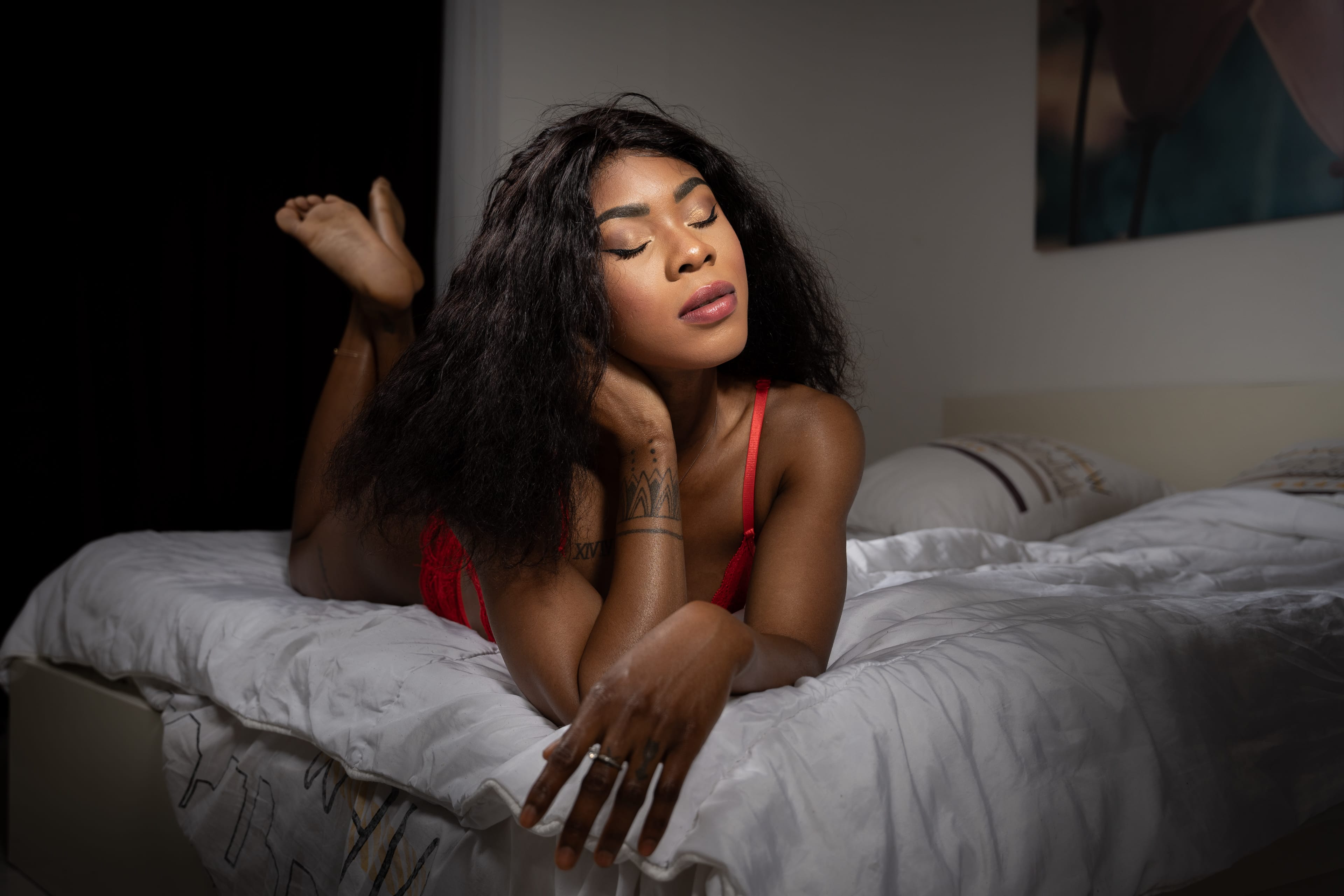 Femme en lingerie rouge allongée sur un lit blanc, les yeux fermés dans un moment serein, une lumière douce mettant en valeur sa présence confiante en boudoir.